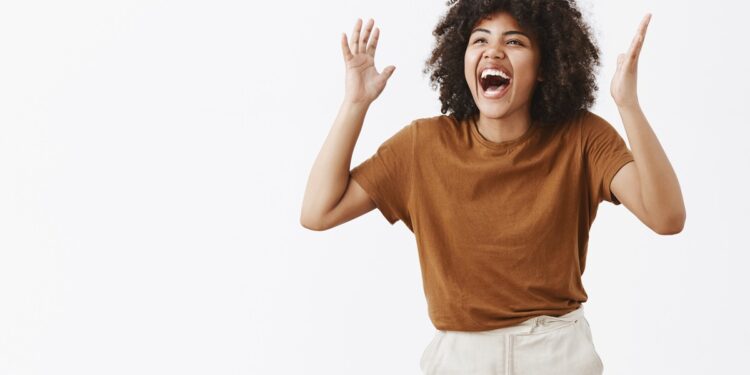 Studio shot of over emotive carefree and happy stylish modern african american female in trendy brown t shrit gesturing with raised hands near head and laughing out loud gazing left