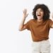 Studio shot of over emotive carefree and happy stylish modern african american female in trendy brown t shrit gesturing with raised hands near head and laughing out loud gazing left