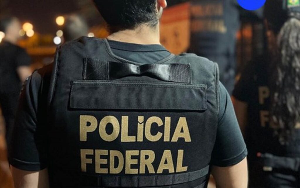 Imagem de um agente da Polícia Federal, com colete preto e a inscrição 'Polícia Federal' nas costas, durante operação noturna.