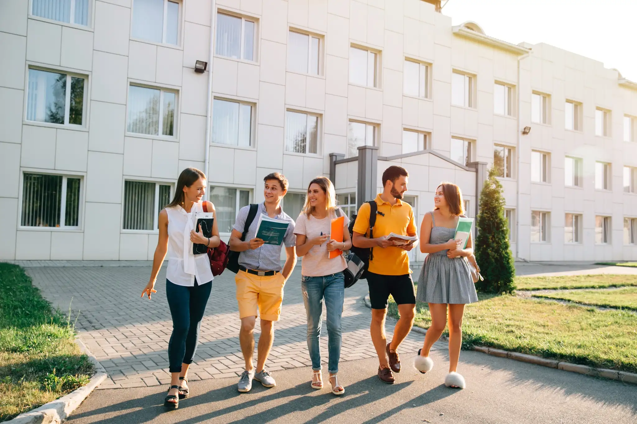 Grupo de jovens caminhando juntos ao ar livre, com livros e pastas, em um ambiente de campus universitário, representando o espírito de preparação para o concurso da Prefeitura de Monte Carmelo.