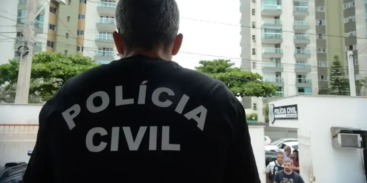 Policial civil com uniforme, com as palavras 'POLÍCIA CIVIL' estampadas nas costas, próximo à entrada da delegacia