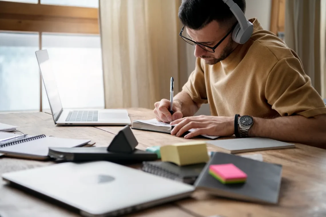 Homem estudando com fones de ouvido, escrevendo em caderno, com laptop e materiais de estudo na mesa