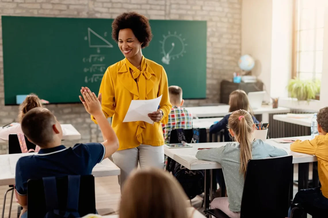 Professora sorrindo e interagindo com alunos em sala de aula, com quadro negro ao fundo