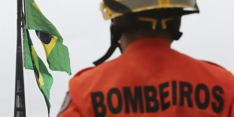 Soldado bombeiro com uniforme laranja olhando para as bandeiras do Brasil ao fundo