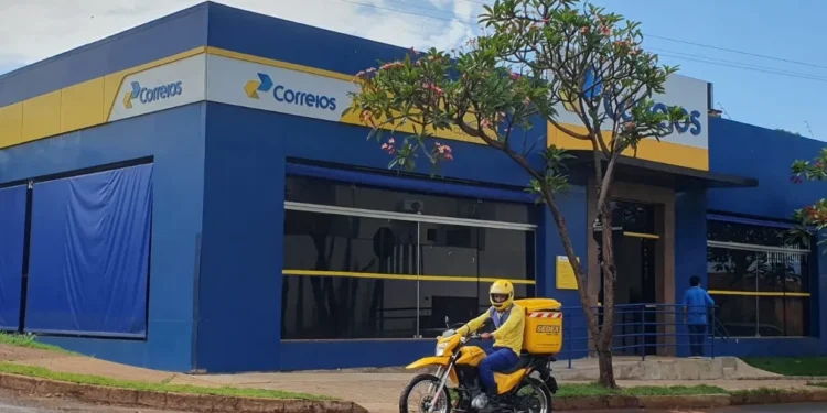 Fachada de uma agência dos Correios com um motociclista entregador passando em frente.