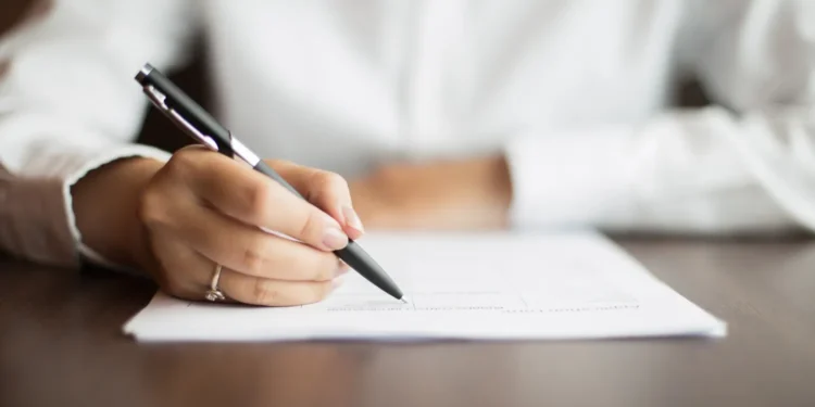 Mão segurando uma caneta e escrevendo em um documento sobre uma mesa