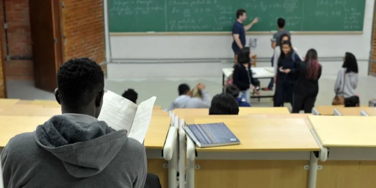  Estudante em sala de aula focado no estudo enquanto outros conversam ao fundo.