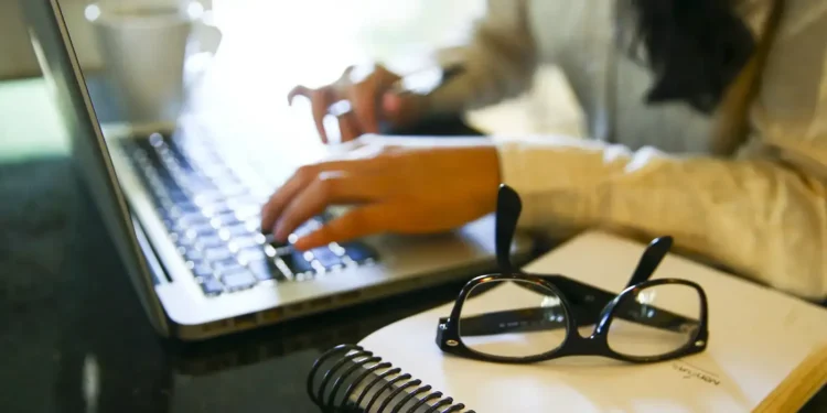 Pessoa digitando no laptop, com óculos e um caderno em primeiro plano.