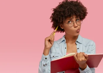 Mulher com cabelo cacheado e óculos, segurando um caderno e pensando, representando a dúvida comum sobre a grafia correta de palavras