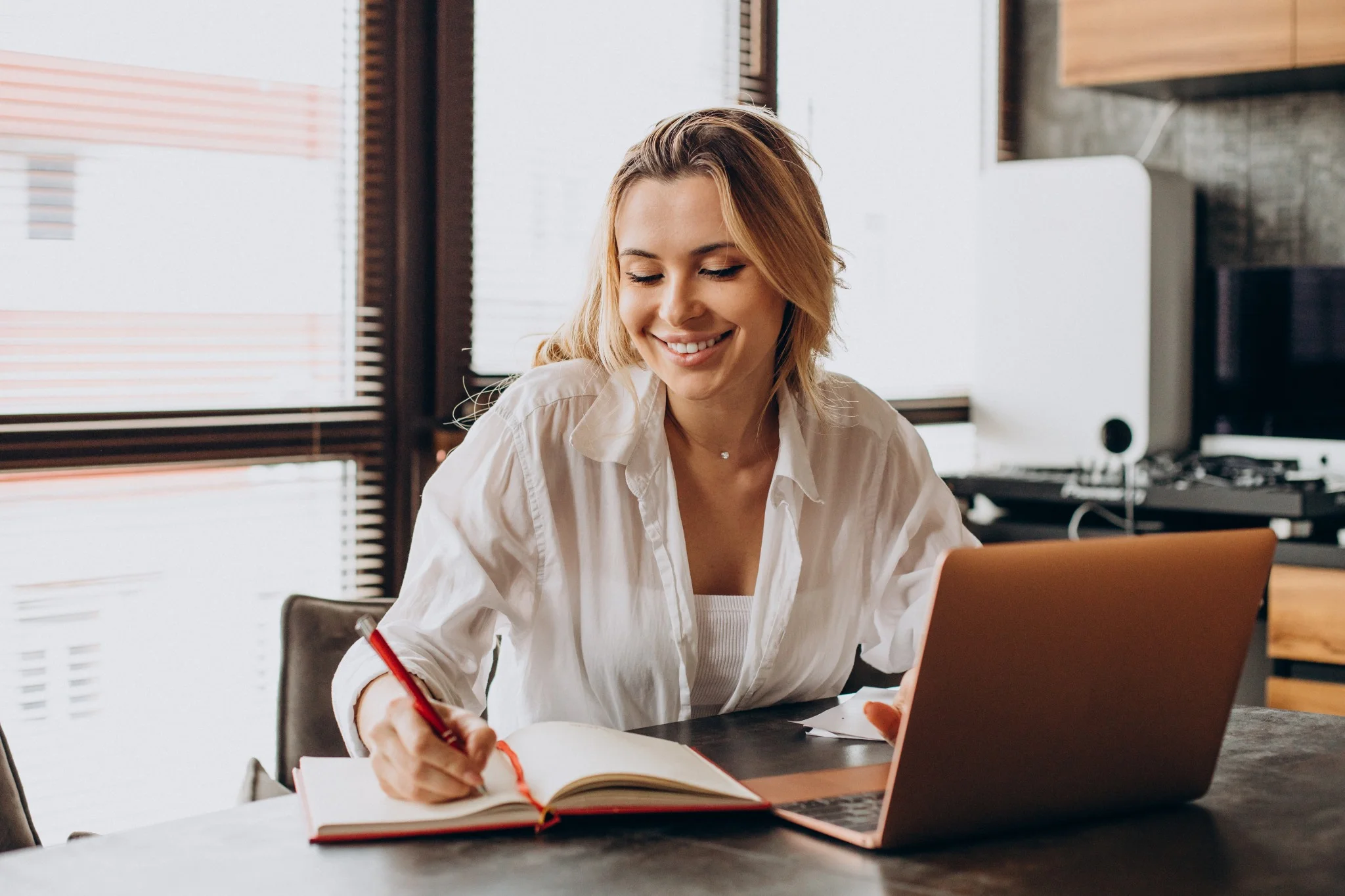 Mulher sorrindo enquanto trabalha de casa, com um laptop e um caderno aberto, representando o estudo para concursos.