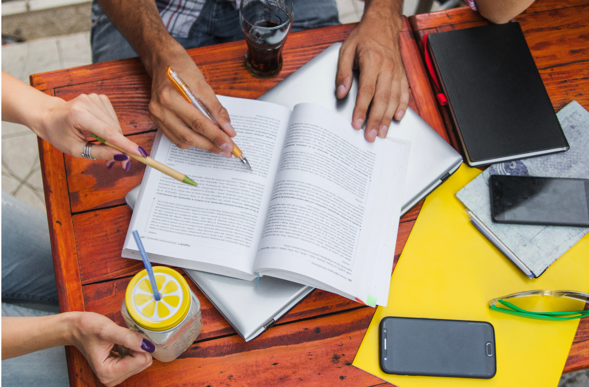 Grupo de estudantes revisando conteúdos e fazendo anotações em um livro aberto, com materiais de estudo organizados sobre a mesa.