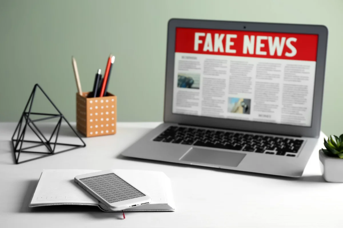 Imagem de um laptop com uma tela exibindo o título 'FAKE NEWS', ao lado de um caderno e um smartphone, simbolizando a disseminação de notícias falsas.