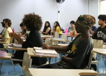 Estudantes em uma sala de aula, atentos durante uma aula, com foco em um aluno escrevendo.