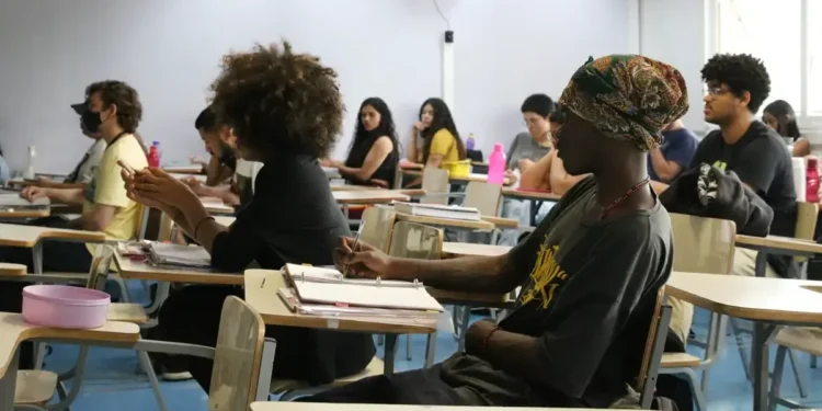 Estudantes em uma sala de aula, atentos durante uma aula, com foco em um aluno escrevendo.