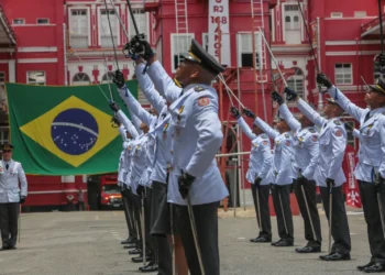 Militares do Corpo de Bombeiros do Rio de Janeiro realizam cerimônia com bandeira do Brasil ao fundo.