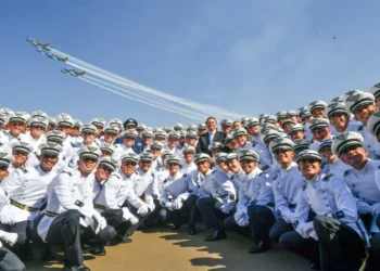 Grupo de cadetes da Aeronáutica sorrindo em cerimônia oficial, com esquadrilha da fumaça ao fundo