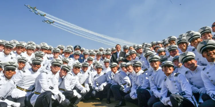 Grupo de cadetes da Aeronáutica sorrindo em cerimônia oficial, com esquadrilha da fumaça ao fundo