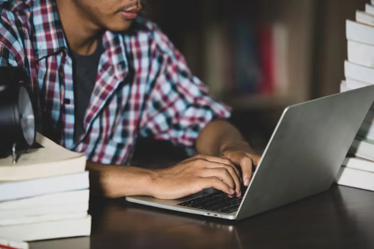 Jovem estudando para concurso no computador, com livros ao redor.