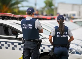 Dois policiais militares observando a patrulha, com viaturas ao fundo.
