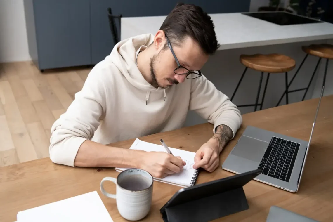 Homem estudando para concurso público, escrevendo em um caderno com laptop e tablet sobre a mesa