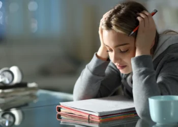 Jovem estudante com dificuldades durante os estudos, com a cabeça apoiada nas mãos e um livro aberto à sua frente.