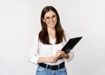Mulher de óculos sorrindo e segurando uma prancheta, com camisa branca e jeans, sobre fundo neutro.