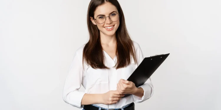 Mulher de óculos sorrindo e segurando uma prancheta, com camisa branca e jeans, sobre fundo neutro.