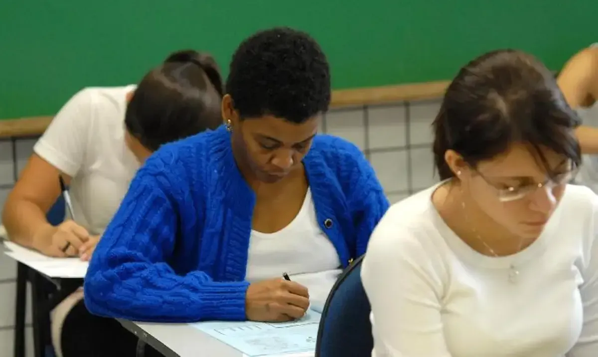 Estudantes em sala de aula durante um exame, com foco em uma mulher de cabelo curto usando um suéter azul.
