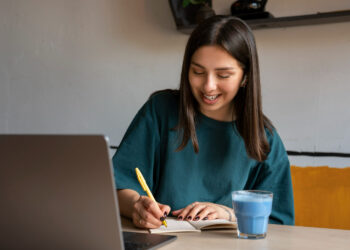 Jovem sorridente estudando em casa com caderno, caneta amarela, laptop aberto e copo de bebida azul.