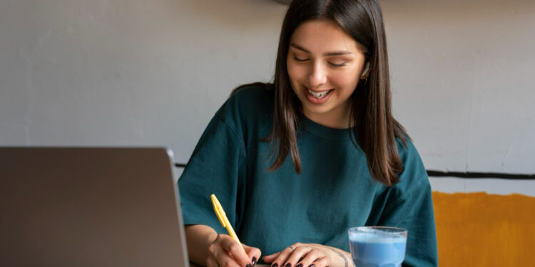 Jovem sorridente estudando em casa com caderno, caneta amarela, laptop aberto e copo de bebida azul.
