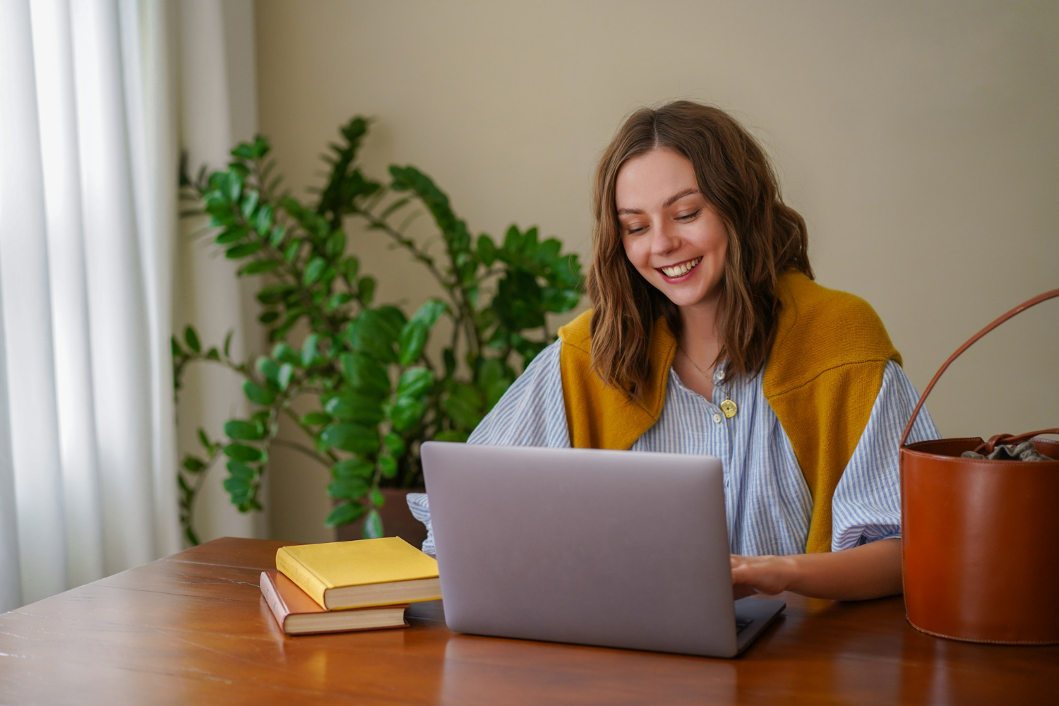 Mulher sorridente estudando no laptop em uma mesa com livros e planta ao fundo.