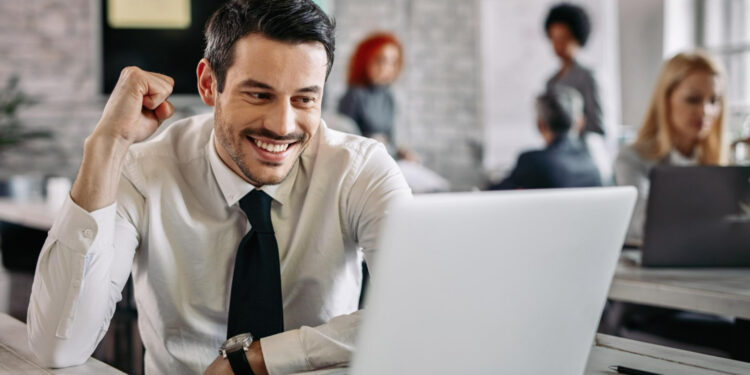 Homem sorridente em ambiente de escritório comemorando sucesso ao olhar para o laptop, com colegas de trabalho desfocados ao fundo.