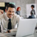 Homem sorridente em ambiente de escritório comemorando sucesso ao olhar para o laptop, com colegas de trabalho desfocados ao fundo.