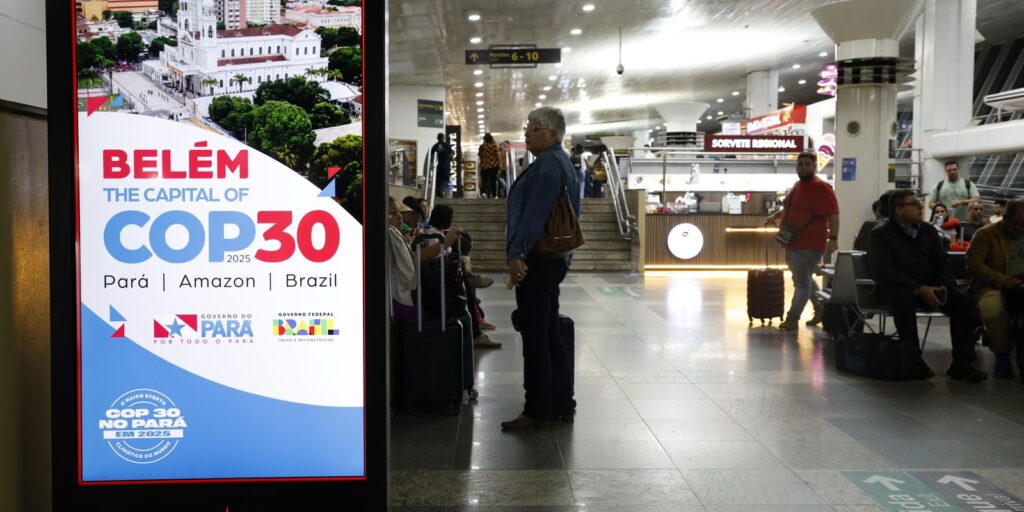 Painel informativo da COP 30 em Belém com pessoas em terminal de aeroporto ao fundo