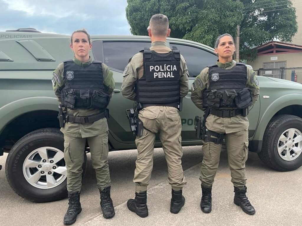 Três policiais penais em uniforme e colete tático, em frente a veículo oficial verde.