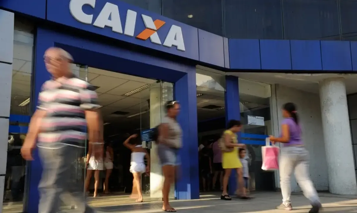 Entrada movimentada da agência Caixa com pessoas passando e fila na porta.