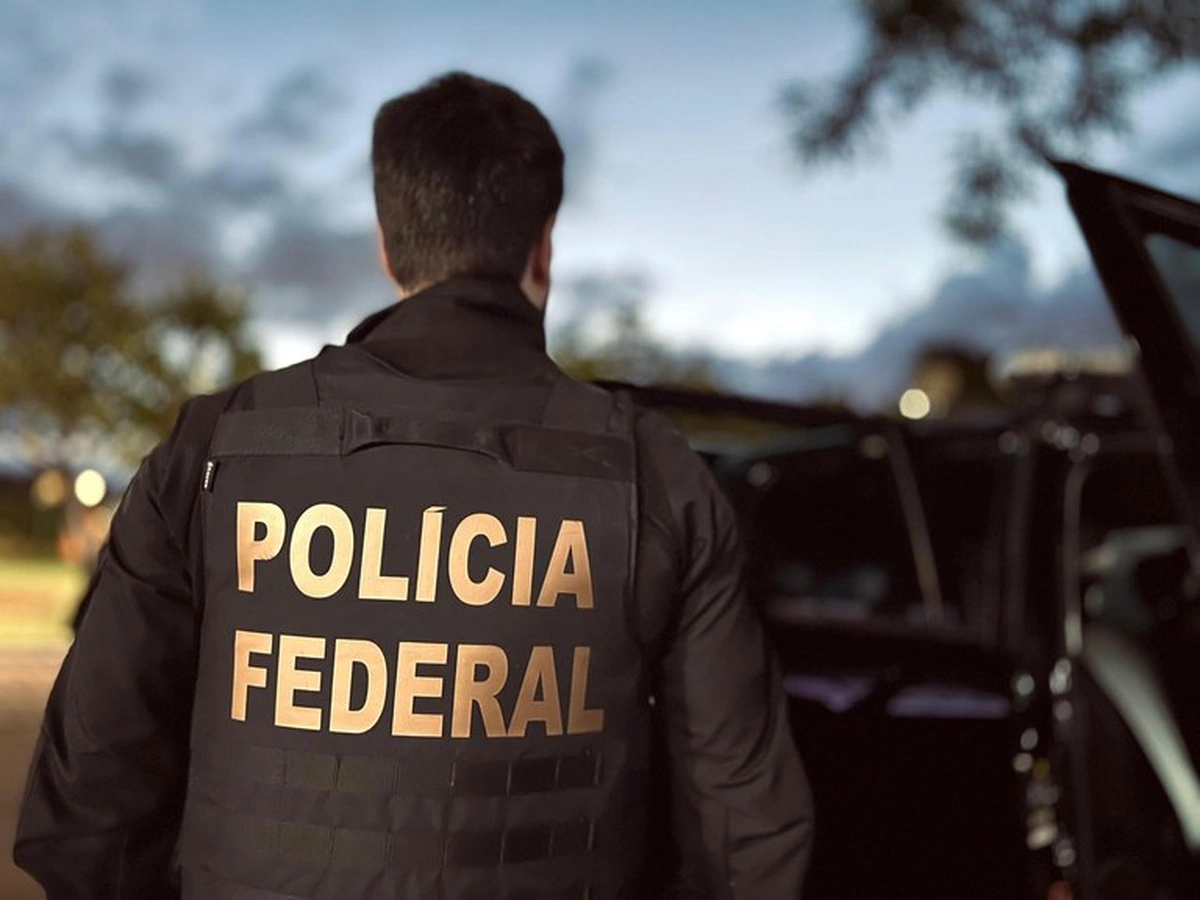 Agente da Polícia Federal usando colete tático, visto de costas, durante operação à noite.