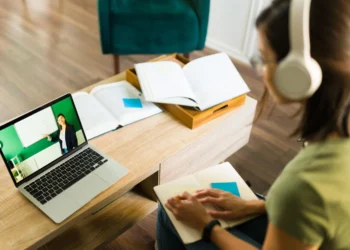 Estudante participa de aula online usando fones de ouvido, assistindo a professora explicando conteúdos em quadro branco no computador