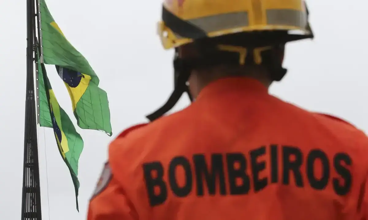 Bombeiro de costas, com uniforme, observando as bandeiras do Brasil ao fundo.