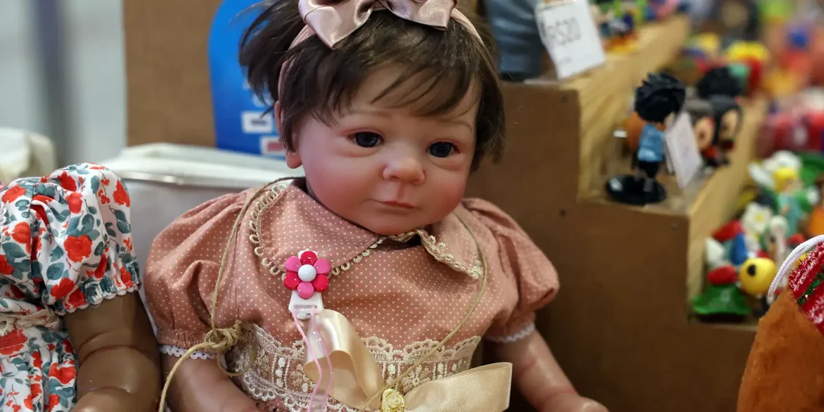 Boneca bebê reborn com vestido rosa e laço, exposta em feira de artesanato.