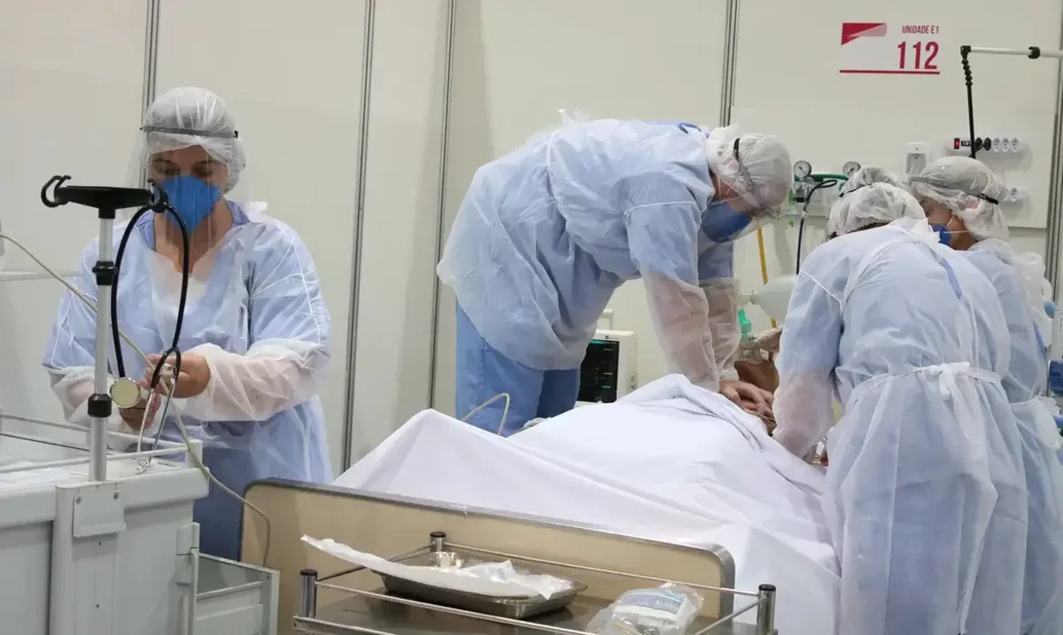 Profissionais de saúde cuidando de um paciente em uma unidade de atendimento em hospital de campanha, com equipamentos de proteção individual.