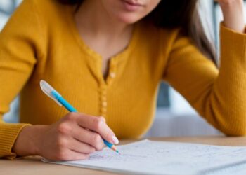 Mulher concentrada escrevendo em papel com caneta azul em mesa clara, usando suéter mostarda.