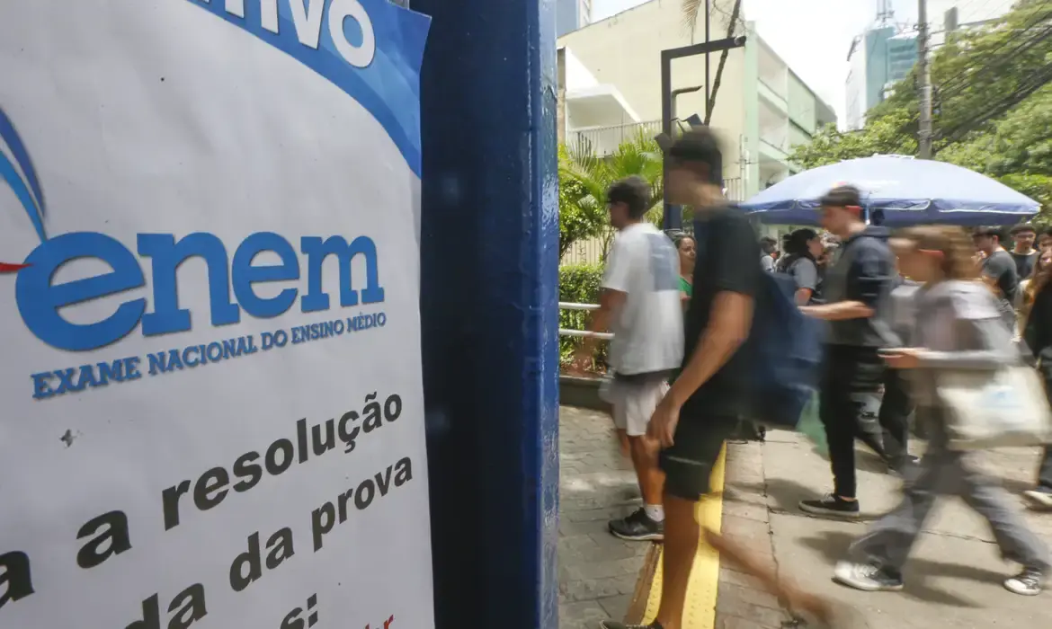 Jovens caminhando em frente a placa do ENEM, Exame Nacional do Ensino Médio, em ambiente urbano.