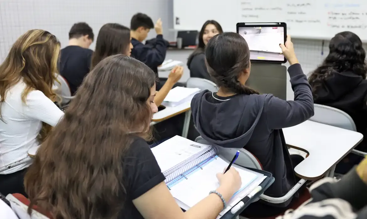 Alunos estudando em sala de aula, utilizando cadernos e tablets.