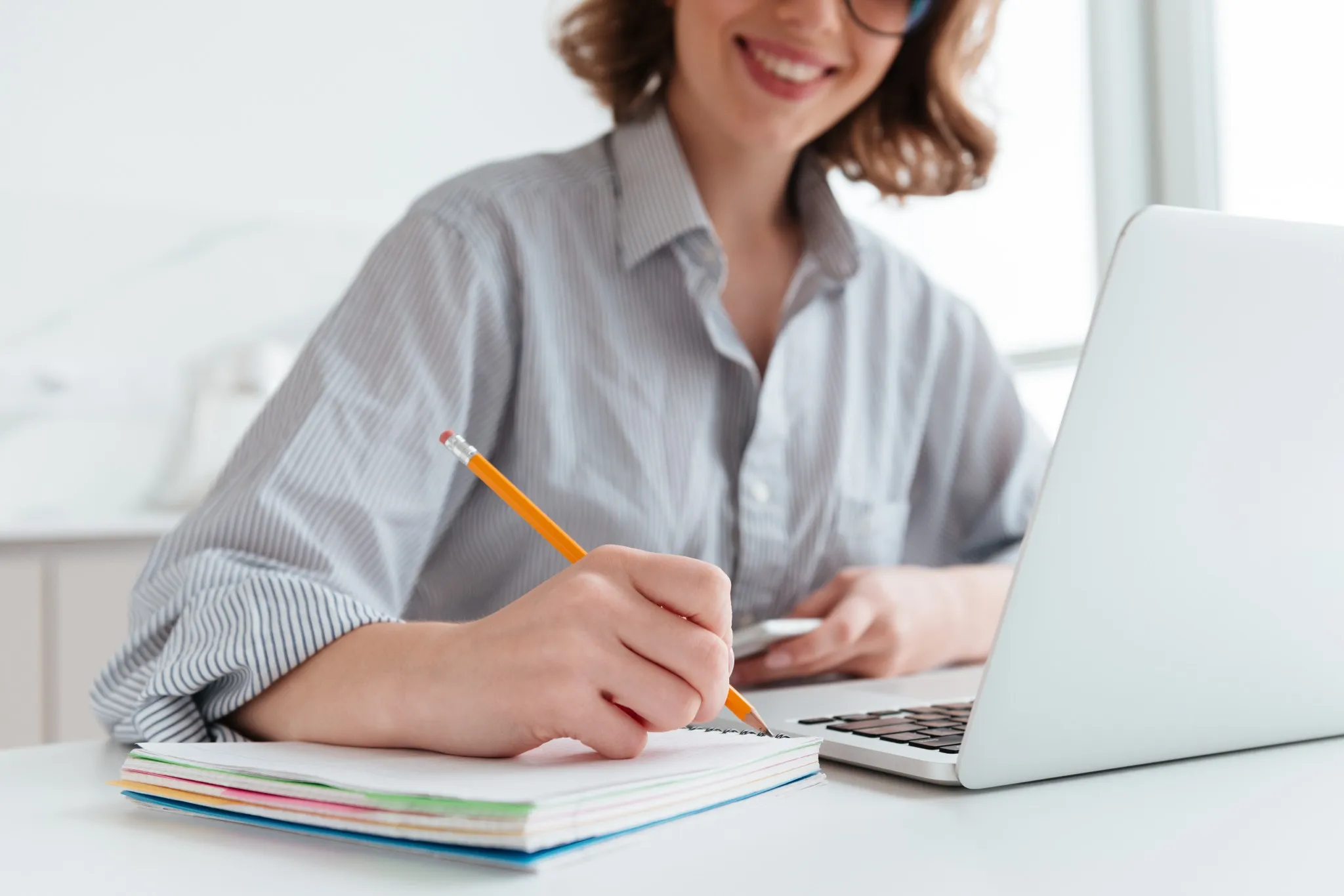 Jovem mulher sorridente usando camisa listrada, escrevendo em caderno ao lado de um laptop aberto.