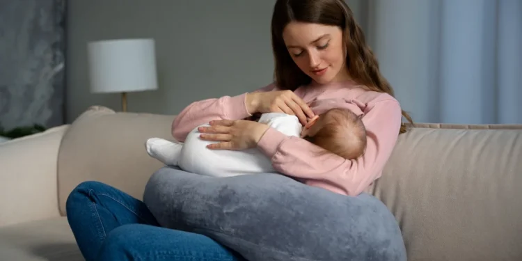 Mãe amamentando bebê recém-nascido com apoio de almofada no sofá.