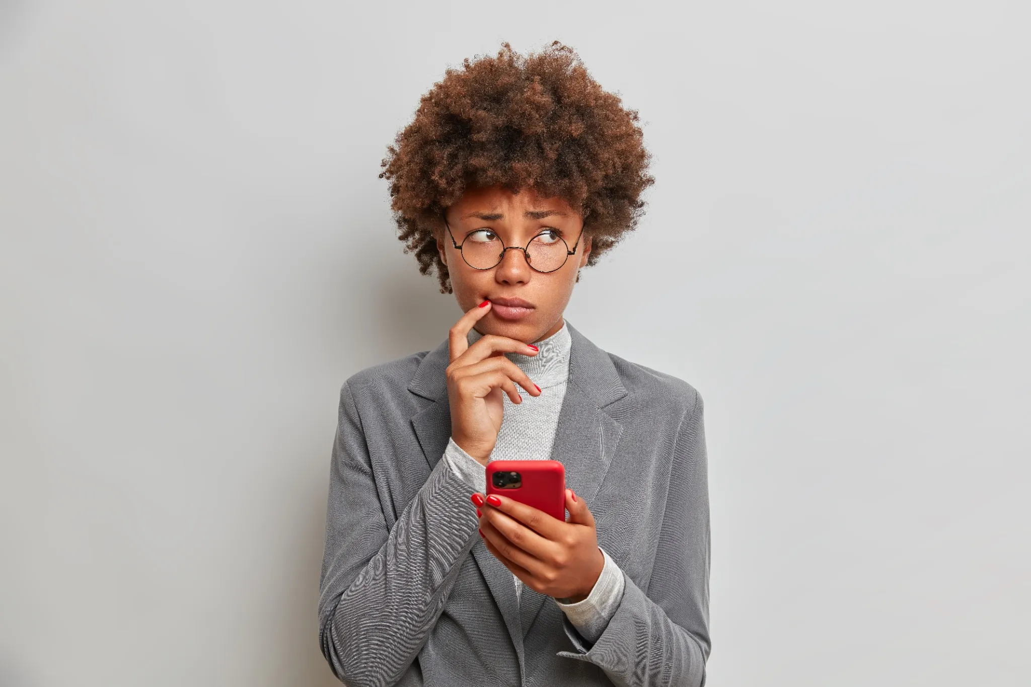 Mulher jovem pensativa usando óculos redondos segurando celular vermelho.