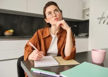 Jovem mulher pensativa escrevendo em um caderno com caneta rosa na cozinha moderna