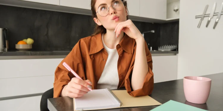 Jovem mulher pensativa escrevendo em um caderno com caneta rosa na cozinha moderna