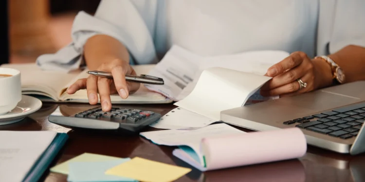Pessoa calculando despesas financeiras com calculadora, notas fiscais e laptop sobre mesa.